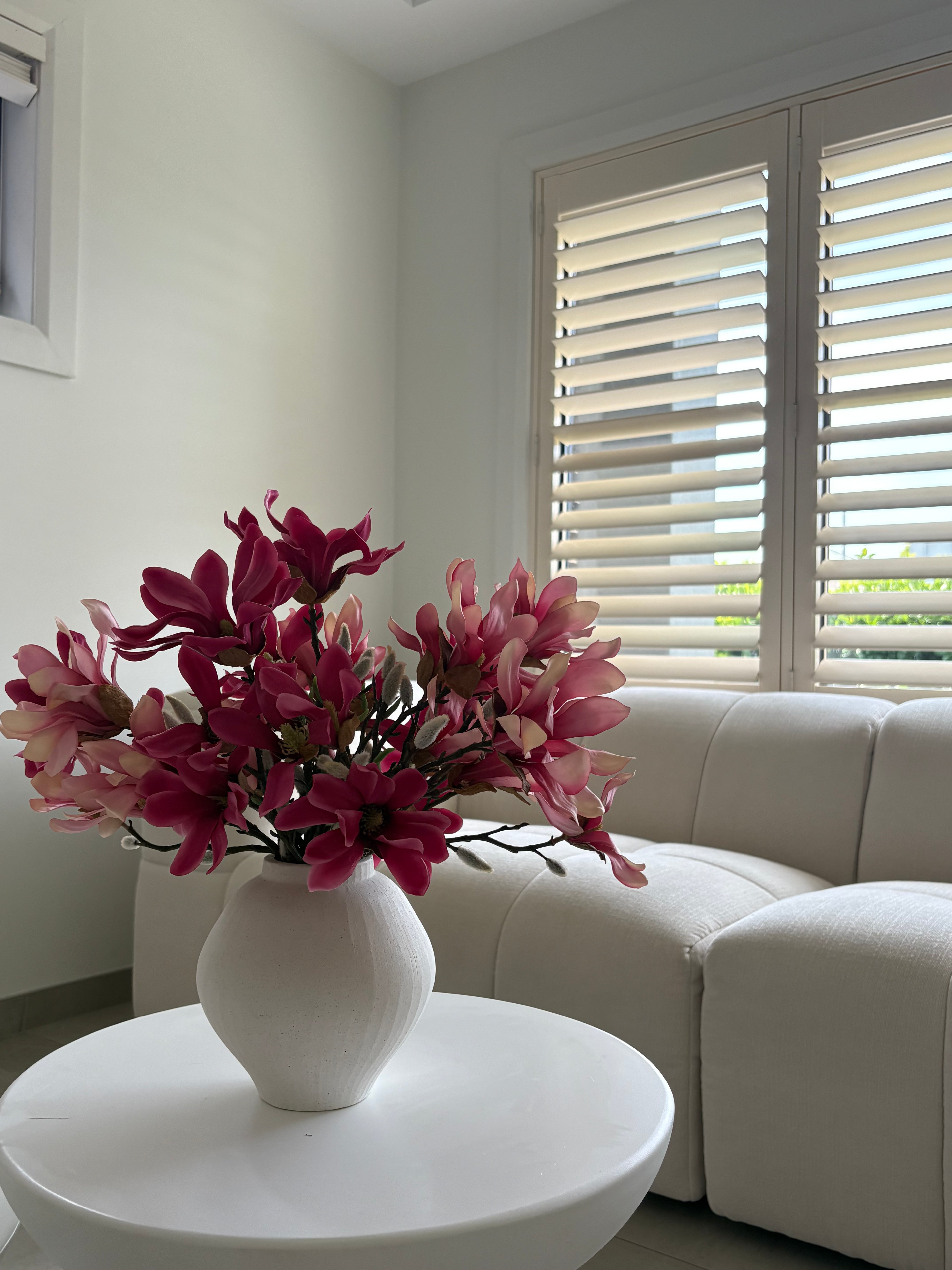 A white ceramic vase filled with pink and magenta artificial flowers, placed on a round white side table in a bright living room with a cream sofa and window shutters.