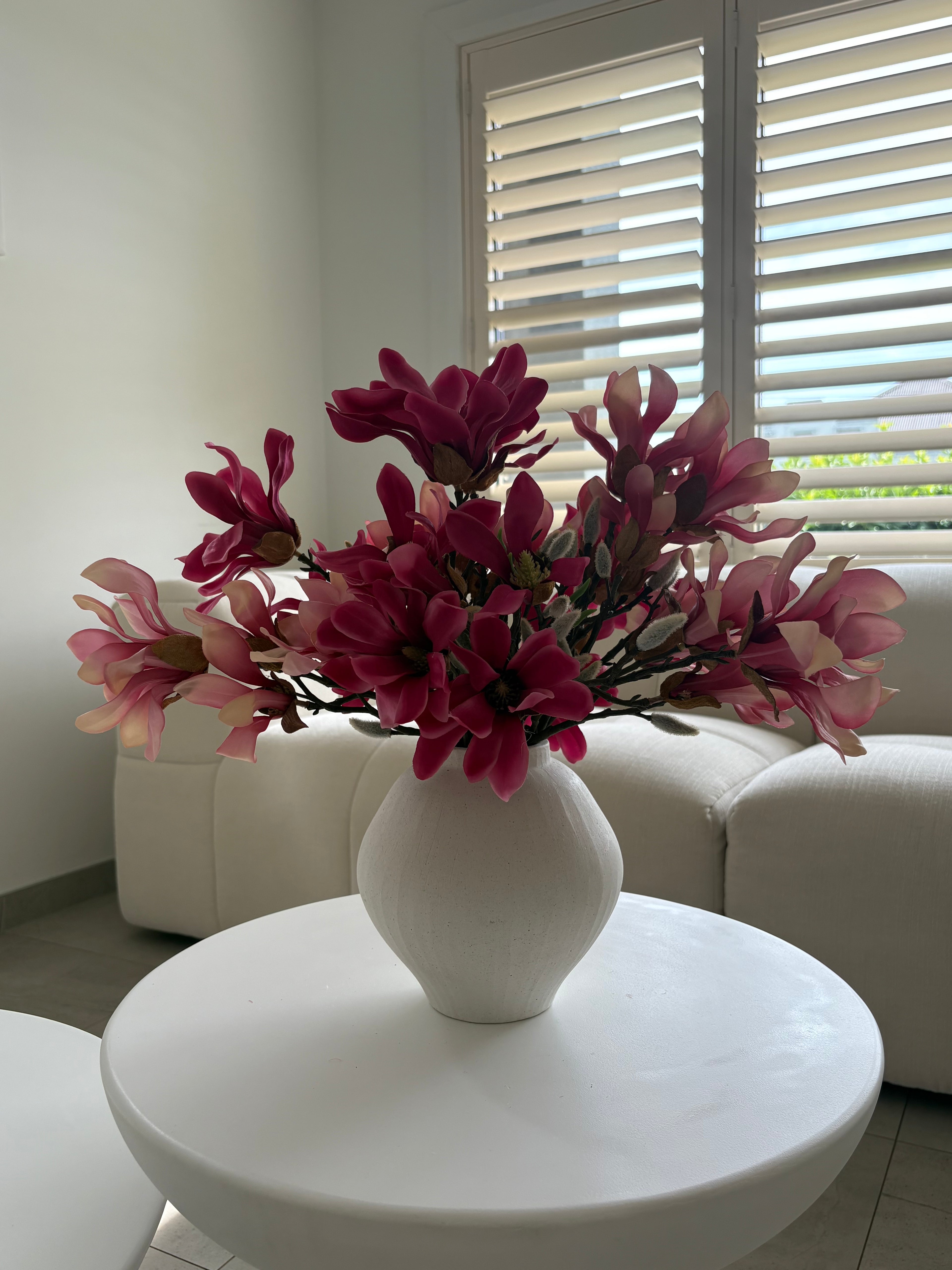 A white ceramic vase filled with artificial pink and magenta magnolia flowers, placed on a round white table in a bright living room with a white sofa and window blinds in the background.
