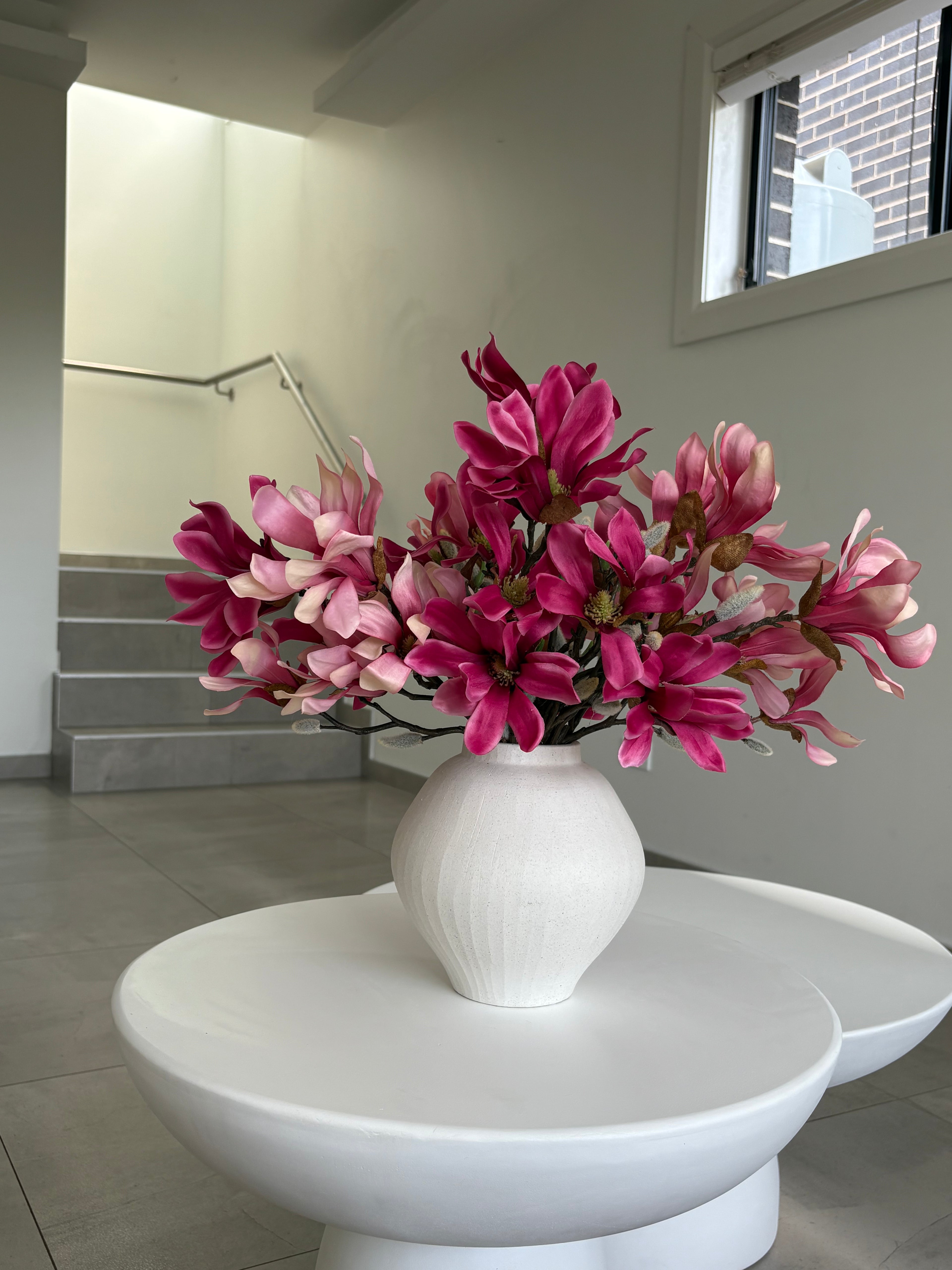A white ceramic vase filled with vibrant pink and magenta magnolia flowers, displayed on a modern white sculptural pedestal table in a minimalist interior setting.