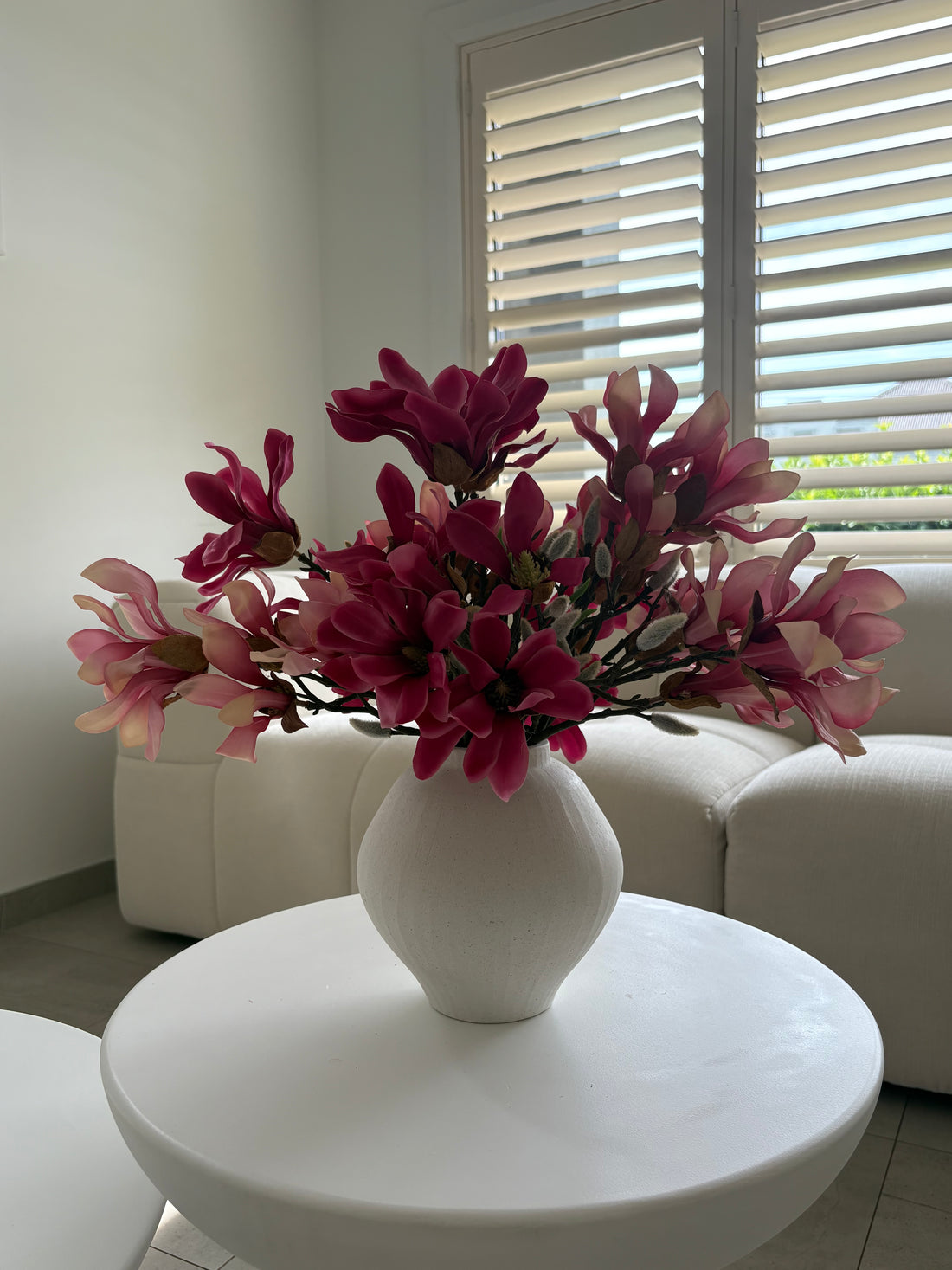 A white ceramic vase filled with artificial pink and magenta magnolia flowers, placed on a round white table in a bright living room with a white sofa and window blinds in the background.