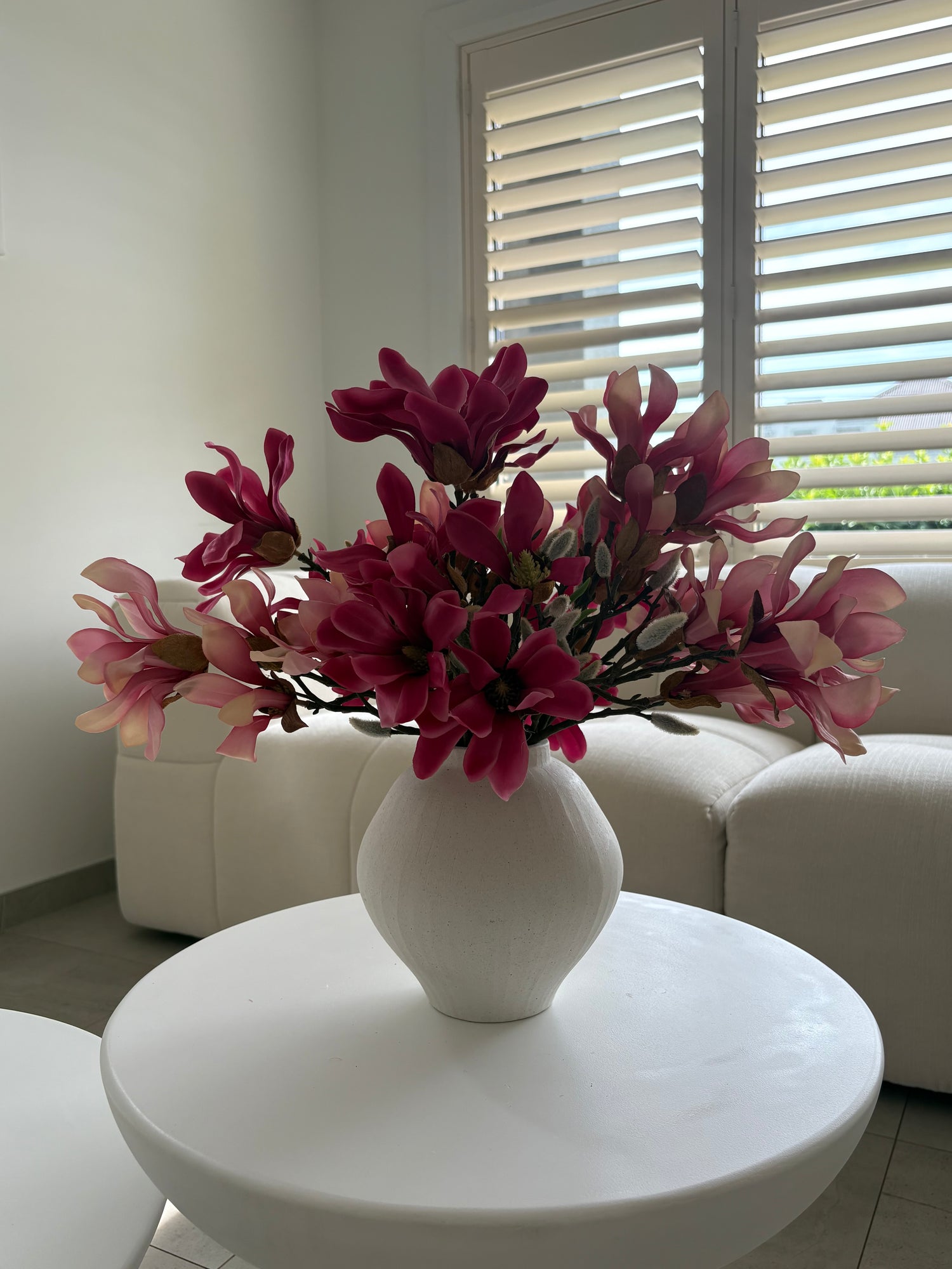 A white ceramic vase filled with artificial pink and magenta magnolia flowers, placed on a round white table in a bright living room with a white sofa and window blinds in the background.