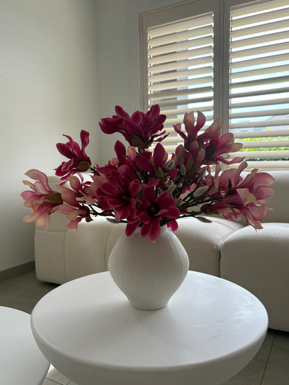 A white ceramic vase filled with artificial pink and magenta magnolia flowers, placed on a round white table in a bright living room with a white sofa and window blinds in the background.